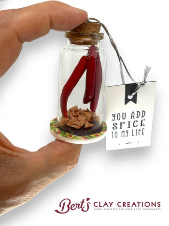 Miniature bottle with red chili peppers and clay tortilla chips, held by a hand, with a 'You Add Spice to My Life' tag and 'Bert's Clay Creations' branding.